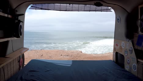 Travel-van-with-back-door-open-to-ocean-view-with-surfers-in-the-distance,-Inside-shot