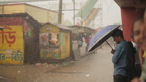 Young-Indian-people-stuck-in-rain,-male-waiting-in-rain-with-umbrella-open-looking-outside,-monsoon-season,-slow-motion-shot-of-rain-drops