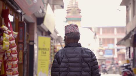 Zeitlupe-Von-Hinten-Eines-Nepalesischen-Alten-Mannes-Mit-Traditionellem-Hut,-Der-Auf-Der-Straße-Läuft,-Mit-Einem-Tempel-Im-Hintergrund,-Kathamdu,-Nepal