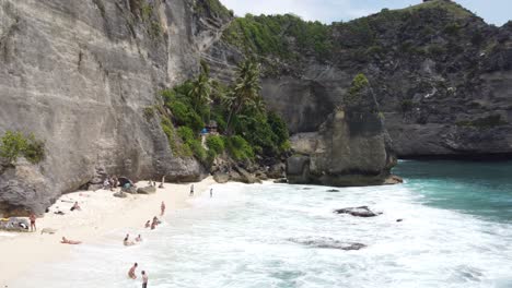 Aerial,-Diamond-Beach-in-Nusa-Penida-island,-Tourists-people-swinging,-sunbathing,-and-swimming-in-tropical-azure-water-on-white-sand-beach-down-a-steep-limestone-cliffside