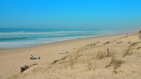 Vista-Panorámica-De-Turistas-Sentados-En-La-Arena-Mientras-Observan-Las-Olas-Del-Mar-Rompiendo-En-La-Orilla-Del-Mar-Durante-El-Día-En-Nueva-Aquitania,-Francia