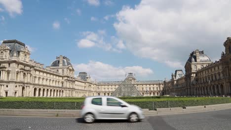 Tráfico-Que-Pasa-Con-El-Patio-De-La-Pirámide-Del-Louvre-Y-El-Palacio-Al-Fondo-En-París,-Francia