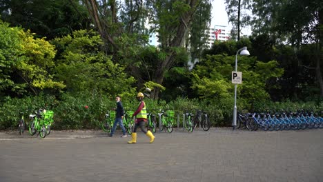 Escena-De-Una-Persona-Estacionada-Con-Las-Bicicletas-Alquiladas-Y-Un-Trabajador-Migrante-Extranjero-Que-Lleva-Un-Objeto-Pasa-Por-El-Espacio-De-Estacionamiento-De-Bicicletas-En-Los-Jardines-Del-Lago-Jurong,-Singapur