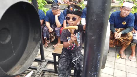 Un-Niño-Pequeño-Toca-El-Instrumento-Musical-Gamelan-Gong-En-Bali-Indonesia-En-La-Ceremonia-De-Cremación,-Hinduismo-Balinés-Con-Gafas