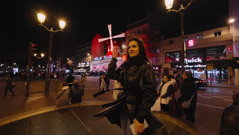 Beautiful-young-woman-smiling-in-front-of-the-famous-Moulin-Rouge-building,-with-red-neon-signs,-in-Paris,-France,-at-night