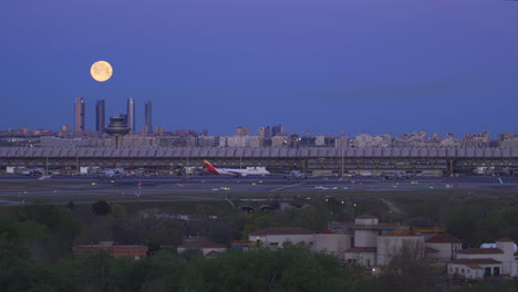 Full-moon-setting-behind-the-Four-Tower-and-International-Airport-in-Madrid,-Spain