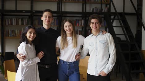 Zoom-out-portrait-of-four-multi-ethnic-young-people-standing-together-in-group-looking-at-camera-posing-at-library-or-in-modern