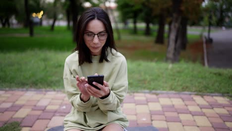 A-brunette-girl-in-a-green-sweater-with-glasses-sits-on-a-skateboard-in-the-park-and-texts-on-the-phone.-Evening-walk,-leisure