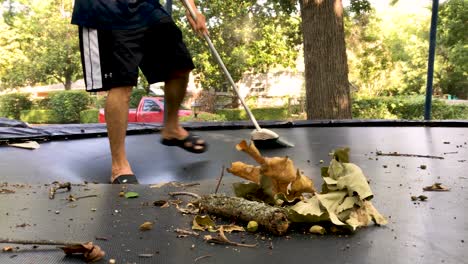 A-real-time-shot-of-a-man-cleaning,-sweeping-the-leaves-from-his-black-coloured-trampoline-in-his-backyard