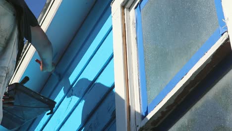 Do-it-yourself-house-painting-in-Waco,-Texas-in-USA,-steady-shot-from-underneath-of-guy-painting-blue-paint-in-slow-motion
