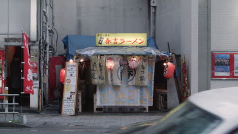 Road-traffic-and-pedestrians-going-past-small-Japanese-shop-on-street-in-Nagasaki,-Japan