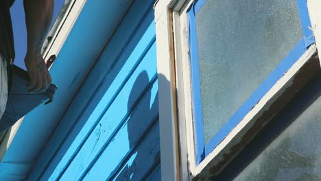 Do-it-yourself-house-painting-in-Waco,-Texas-in-USA,-steady-full-shot-from-the-bottom-of-guy-painting-blue-colour-on-wooden-house