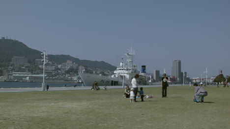 Family-relaxing-on-green-grass-with-a-docked-boat-in-Nagasaki-port-in-the-background,-Japan