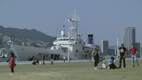 Grupo-De-Adultos-Y-Niños-Sobre-Hierba-Verde-Con-Un-Barco-Atracado-En-El-Puerto-De-Nagasaki-Al-Fondo,-Japón