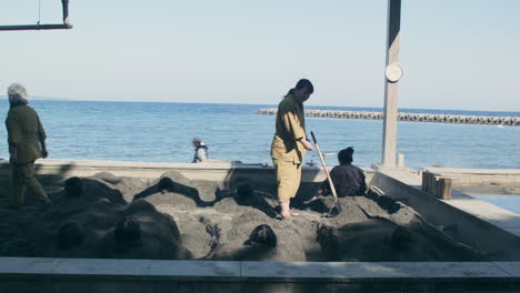 View-of-hot-sand-beach-with-adults-laying-in-them-with-workers-helping-out-and-sea-in-the-background-in-Kagoshima,-Japan