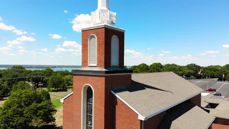 Eine-Drohnenaufnahme-Mit-Aufwärtsbewegung-Des-Sockels-über-Einer-Kirche-Mit-Blick-Auf-Die-Gesamte-Stadt,-Die-Bäume,-Den-See-Und-Die-Straße-Bedeckt,-Mit-Strahlend-Blauem,-Mit-Wolken-Bedecktem-Himmel-Im-Hintergrund