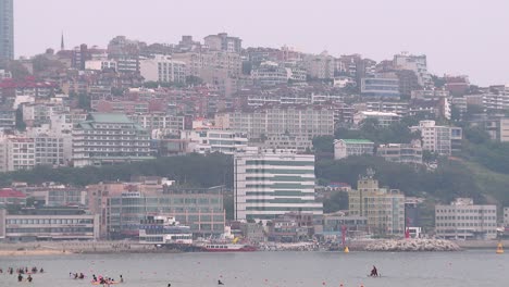 Medium-shot-of-tall-buildings-on-a-hill-next-to-Haeundae-beach-in-South-Korea,-people-playing-in-the-water-and-the-Mipo-Cruise-Terminal-in-the-foreground