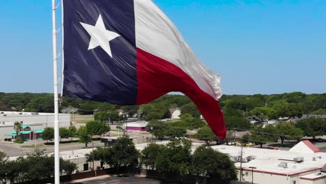 A-close-up-drone-shot,upward-pedestal-motion,-of-the-texas-flag-with-a-bright-blue-sky-and-a-glimpse-of-Wacos-city-in-the-background