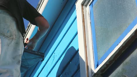 Do-it-yourself-house-painting-in-Waco,-Texas-in-USA,-steady-full-shot-from-the-bottom-of-man-painting-blue-colour-on-wooden-house-in-slow-motion