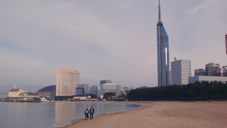 Paar-Geht-Am-Strand-Entlang-Mit-Dem-Fukuoka-Turm-Im-Hintergrund,-Japan