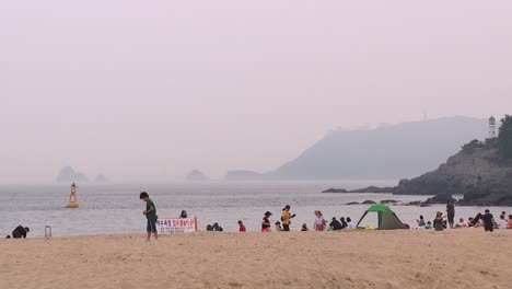 People-having-fun-on-Haeundae-Beach,-mountains-in-the-background,-polluted-sky