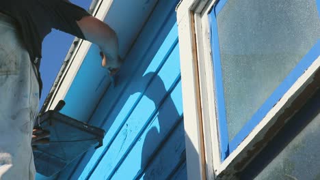 Do-it-yourself-house-painting-in-Waco,-Texas-in-USA,-steady-full-shot-from-the-bottom-of-man-painting-blue-colour-on-wooden-house