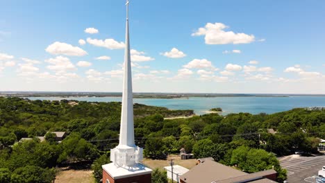 A-drone-shot,-with-upward-pedestal-motion,-over-a-church-with-a-view-of-the-entire-city-covering-trees,lake-and-the-street-with-bright-blue-sky-covered-with-clouds-in-the-background