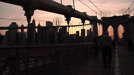 Orangefarbener-Sonnenuntergang-Mit-Silhouette-Der-Brooklyn-Bridge-Mit-Vorbeifahrenden-Menschen-Und-Verkehr-Mit-Der-Skyline-Von-Manhattan-In-Der-Ferne