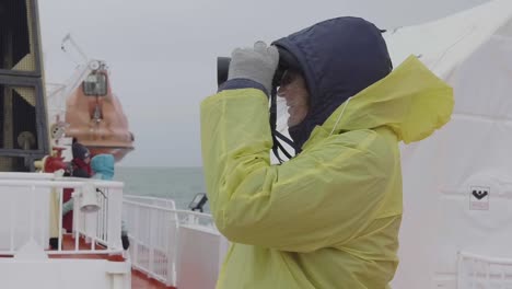 Tourist-Looking-Through-Binoculars-On-Ship-Wearing-Yellow-Raincoat-Off-Greenland