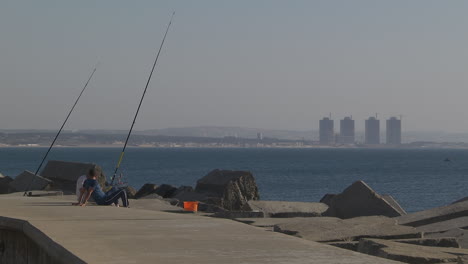 Dos-Pescadores-Sentados-Sobre-Hormigón-Cerca-Del-Mar-Y-Pescando,-Hermoso-Fondo-De-La-Ciudad-De-Argel,-Argelia