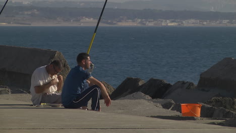 Two-fishermen-sitting-on-concrete-pontoon-and-preparing-bait-for-fishing,-Algiers-city,-Algeria