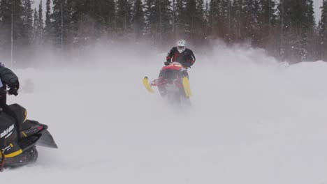 Skidoo-snowmobile-racer-jumps-hill-behind-another-racer-with-jacket-flapping-in-wind-in-snow-motion