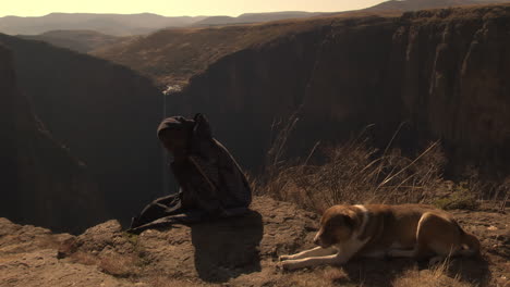 African-woman-and-dog-sitting-on-ground,-Maletsunyane-falls-in-background,-Lesotho,-Africa