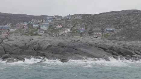 Segeln-Sie-An-Einem-Felsvorsprung-Mit-Tosenden-Wellen-Und-Gebäuden-Vorbei,-Die-Auf-Einem-Hügel-Im-Hintergrund-In-Grönland-Thronen