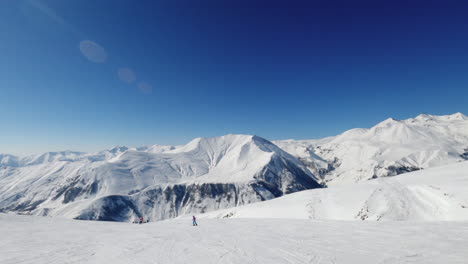 Skiers-ski-down-white-puffy-snow-by-Caucasus-mountains-Gudauri-ski-resort,-Georgia,-pan-left