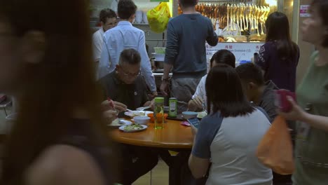 Grupo-De-Personas-Comiendo-Alrededor-De-Una-Mesa-En-Un-Mercado-De-Alimentos-En-Singapur---Toma-Estática-De-Ancho-Medio