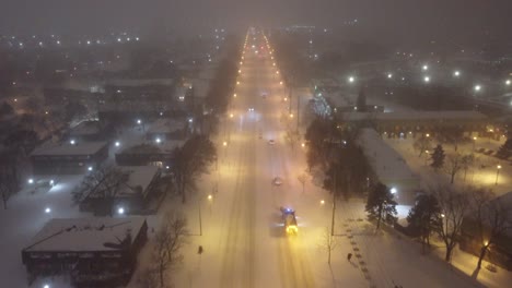 Snow-plow-and-cars-drive-down-suburban-street-covered-in-snow-and-fog-at-night,-aerial-dolly