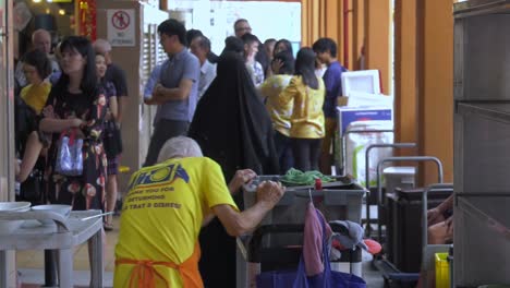 Anciano-Débil-Empujando-Un-Carrito-De-Platos-Sucios-En-El-Centro-De-Alimentos-De-Singapur---Plano-De-Seguimiento-Lento-De-Ancho-Medio