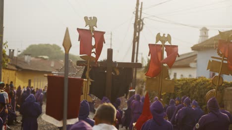 Cucuruchos-En-Túnicas-Moradas-Portando-Carteles-Y-Una-Cruz-De-Madera-En-La-Procesión-De-Semana-Santa-En-La-Calle-De-Antigua-En-Guatemala