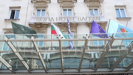 Facade-of-historical-building-of-Sofia-Balkan-Hotel-with-country-flags,-tilt-up