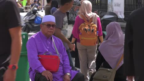 Elderly-Muslim-Male-Sitting-On-Bench-In-Arab-Quarter-With-People-Going-Past