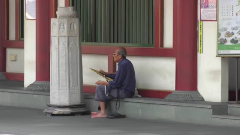 Elderly-man-resting-on-step-refreshing-himself-with-folding-paper-fan---Medium-static-shot