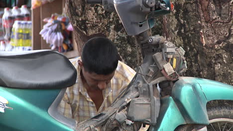 Erwachsener-Mann-Arbeitet-Auf-Einem-Moped-Neben-Einem-Baum-Auf-Der-Straße-In-Iquito,-Peru