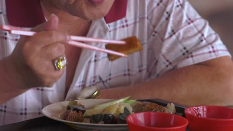 Hombre-Singapurense-De-Mediana-Edad-Comiendo-Carne-De-Cerdo-Y-Comida-Vegetariana-Con-Palillos---Primer-Plano-Inclinado