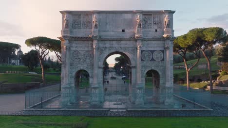 Aerial-drone-view-through-the-sunlit-Arch-of-Constantine,-at-Piazza-del-Colosseo,-sunrise-in-Rome,-Italy
