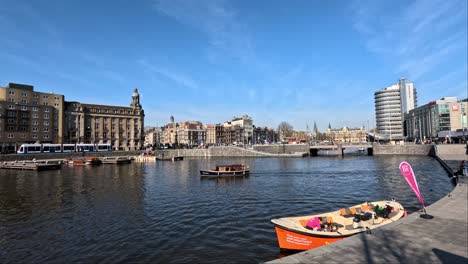 Small-Boat-Sailing-Past-On-Canal-Outside-Amsterdam-Central-Station-On-Sunny-Day-With-Blue-Skies