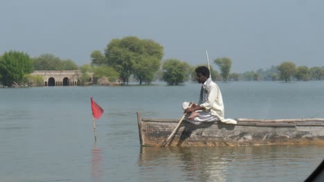 Man-Sitting-On-Forward-Bow-Of-Boat-Slowly-Rowing-On-Flood-Waters-With-Motorbike-Riding-Past-In-Sindh