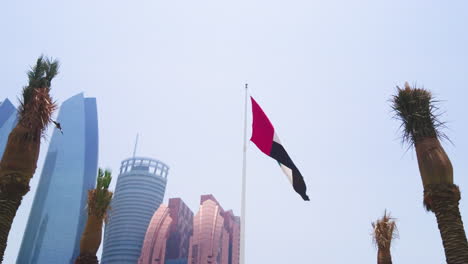 Große-Flagge-Der-Vereinigten-Arabischen-Emirate-Weht-Langsam-Im-Wind-Und-Ein-Falke-Fliegt-Davor,-Eingerahmt-Von-Palmen-In-Der-Mitte,-Mit-Dem-Bab-Al-Qasr-Hotel-Und-Den-Etihad-Towers-Im-Hintergrund-Auf-Der-Corniche-Street-In-Abu-Dhabi,-Nahe-Tagesansicht