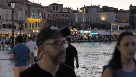 Panning-right-of-people-strolling-at-sunset-along-the-seafront-promenade-with-stores-in-Chania,-Crete