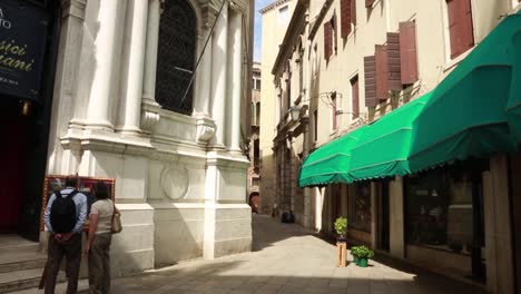 Summer-day-in-Venice-in-a-relatively-empty-street-with-tourist-couple-reading-a-restaurant-menu-board-in-Venice,-Italy